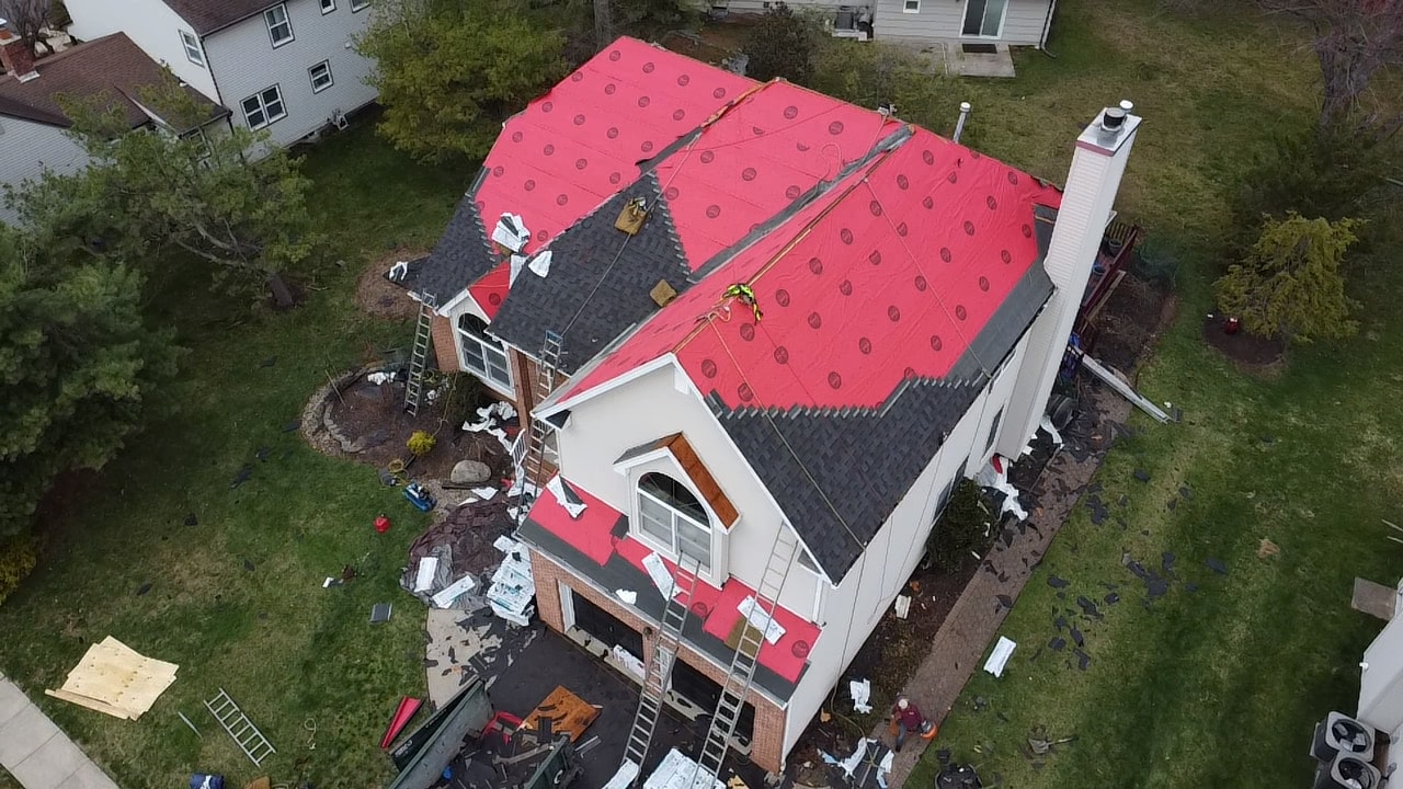 Aerial of a white two-story home with red underlayment on the upper roof and new gray shingles on the lower slopes, crew and ladders visible
