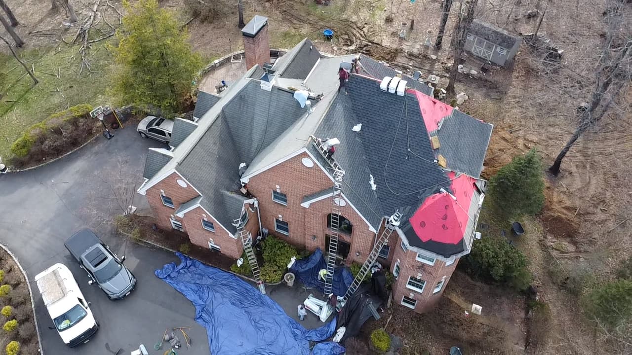 Aerial of a large brick and stucco home mid re-roof, red underlayment over the turret, new gray shingles on other slopes, crew and ladders present